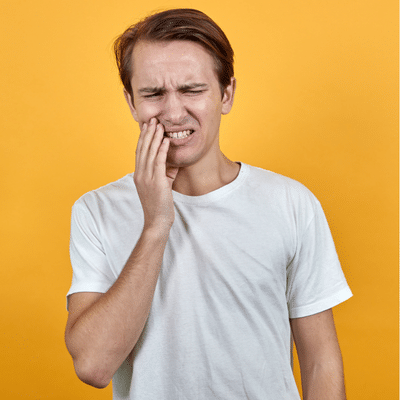 Man wearing white t-shirt holding jaw in pain on a yellow background, representing how smoking affects oral health